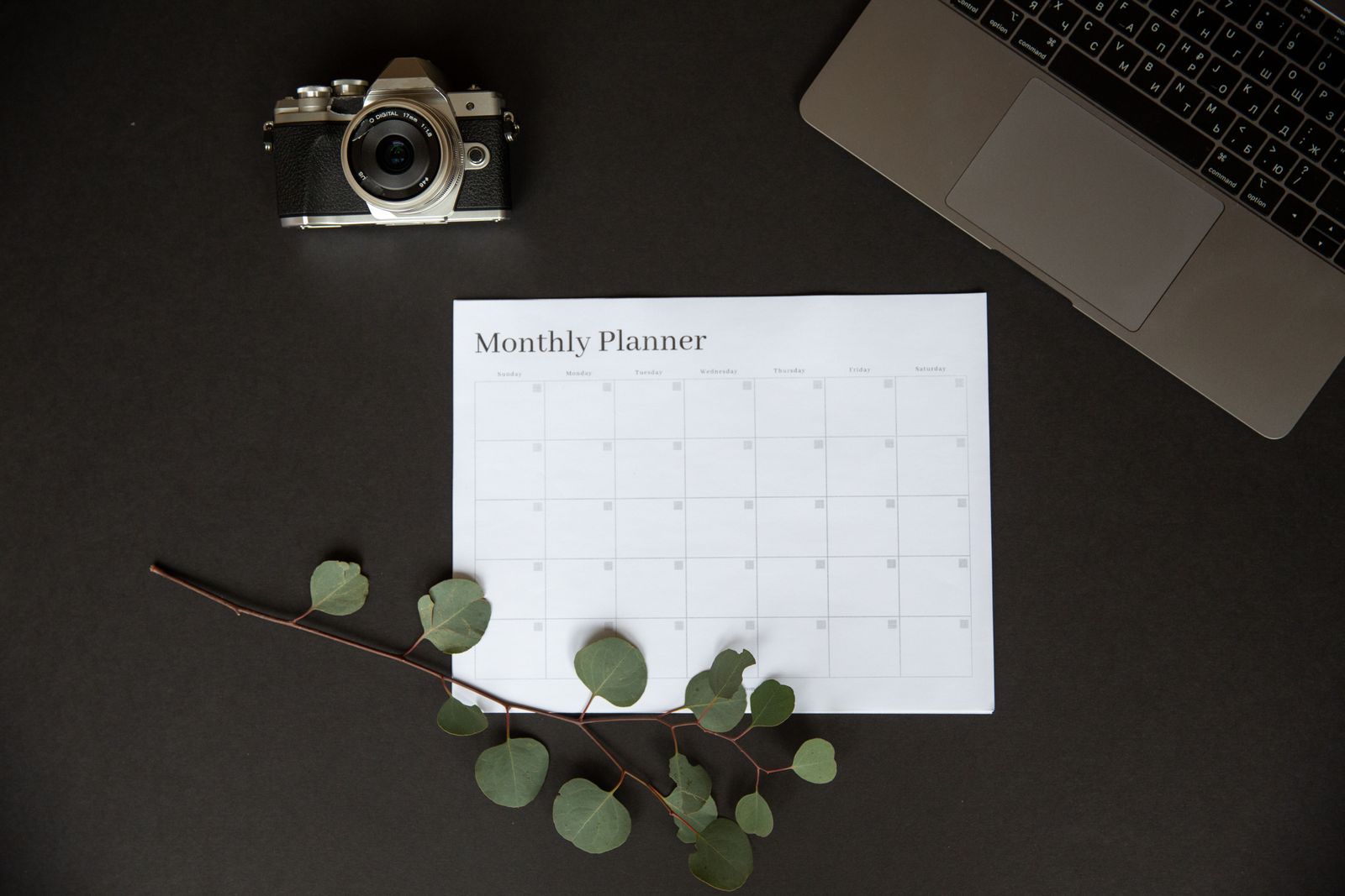 Planner mensal em branco sobre a mesa, ao lado de uma câmera fotográfica e de um notebook