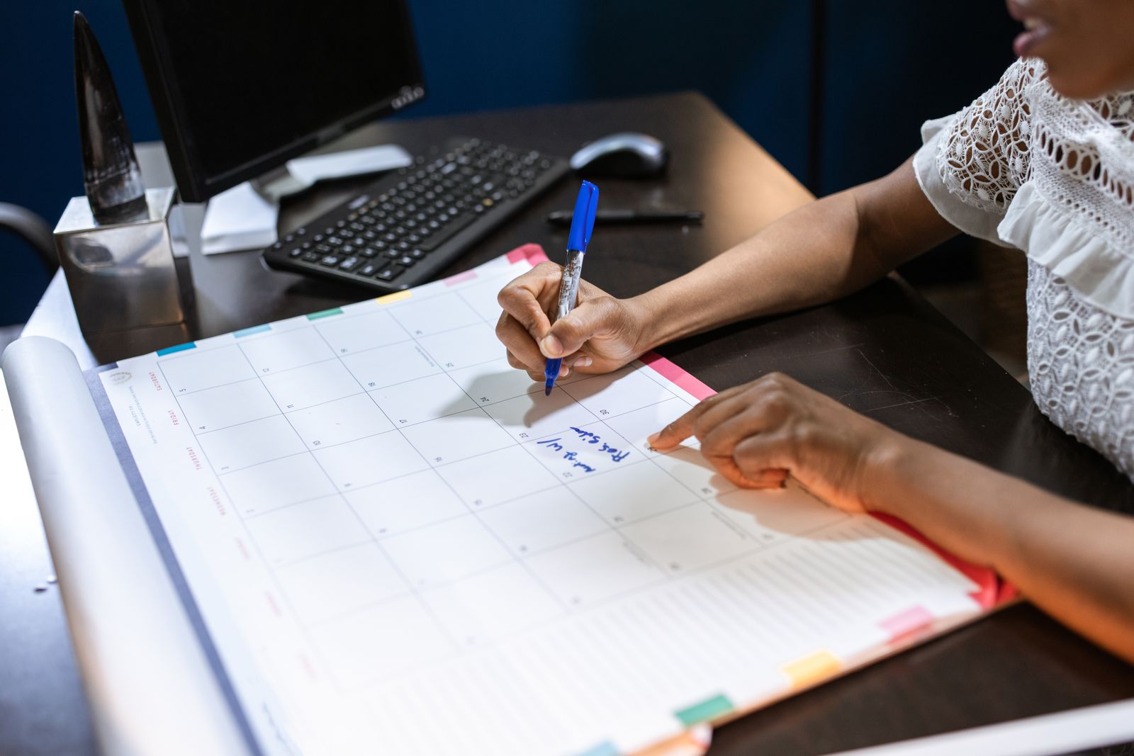 Pessoa marcando um compromisso com caneta num planner de mesa, ao lado do computador