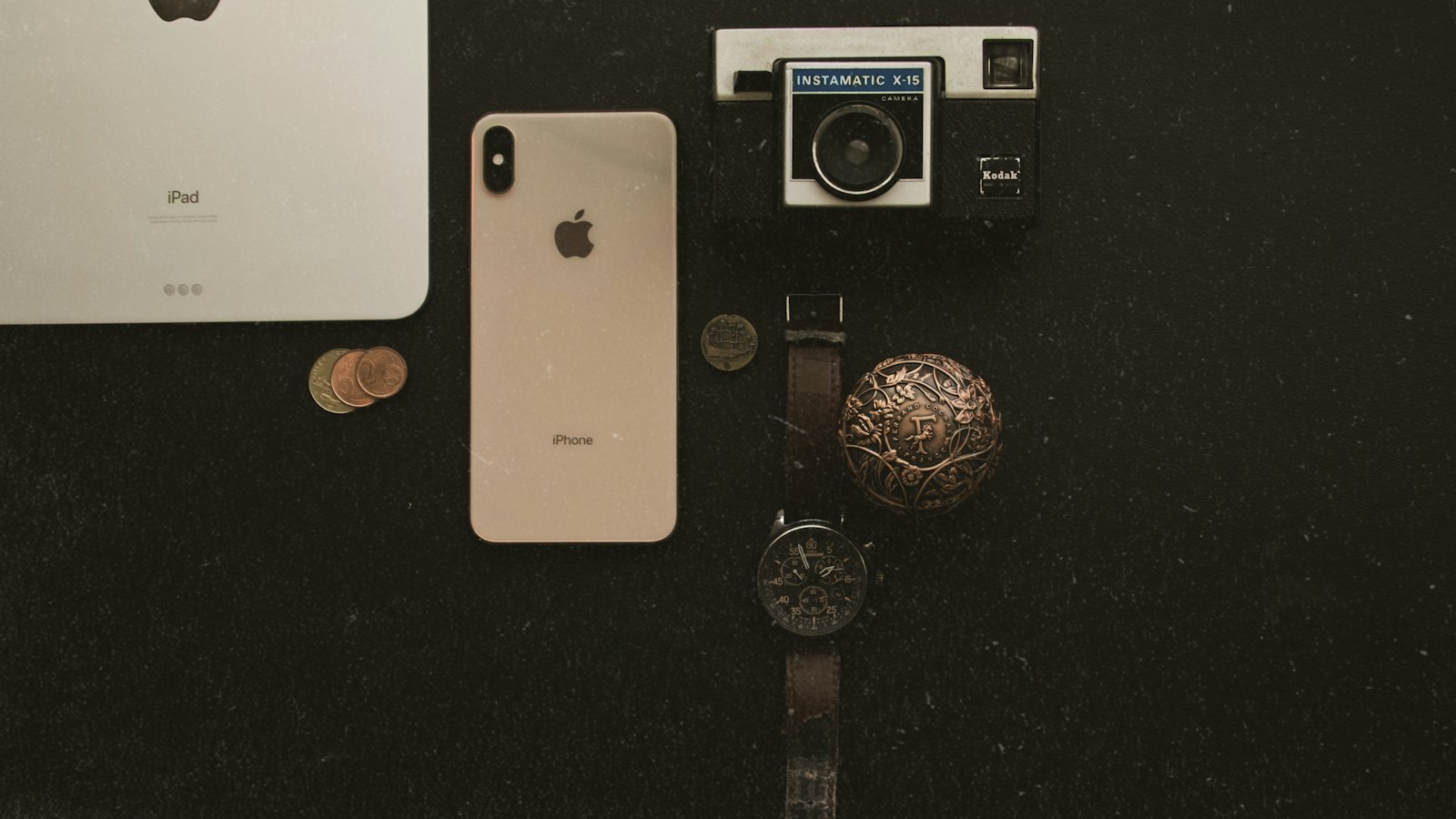 Câmera Kodak antiga ao lado de um iPhone, um iPad e um relógio sobre uma mesa escura, contrastando o equipamento de décadas atrás com o de hoje